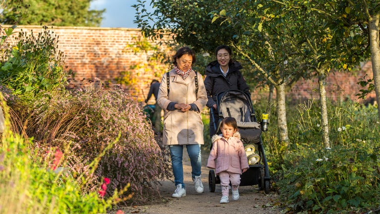 Three family members, including a little girl in a pink coat walking along a path. Trees line the path on one side, and shrubs and flowers on the otehr side. In the background is a red brick wall.